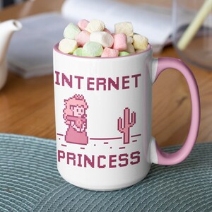 K&ouml;nnte beinhalten: Wei&szlig;er Keramikbecher mit rosa Griff und Rand, gef&uuml;llt mit Marshmallows. Der Becher zeigt pixeligen Text und Grafiken mit der Aufschrift "INTERNET PRINCESS" mit einem Prinzessinnen- und Kaktus-Design.