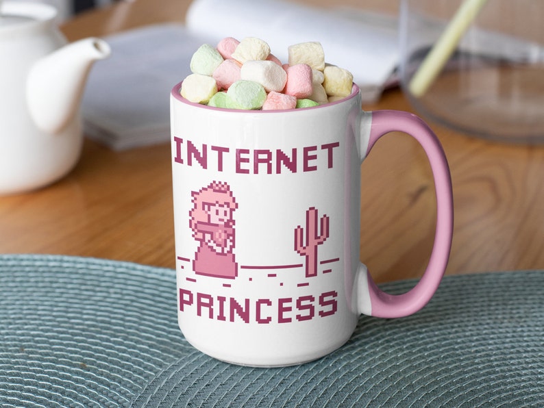 K&ouml;nnte beinhalten: Wei&szlig;er Keramikbecher mit rosa Griff und Rand, gef&uuml;llt mit Marshmallows. Der Becher zeigt pixeligen Text und Grafiken mit der Aufschrift "INTERNET PRINCESS" mit einem Prinzessinnen- und Kaktus-Design.