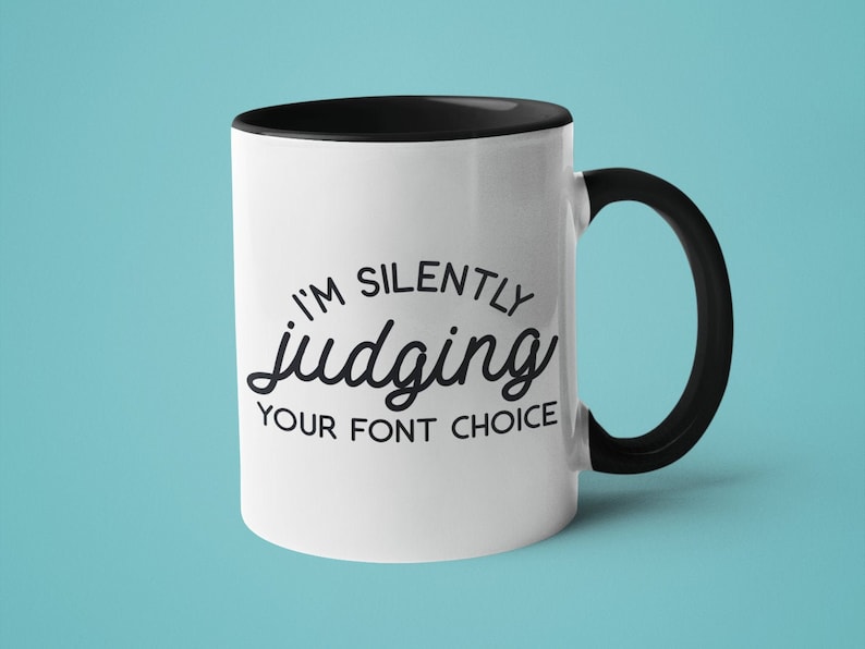 Puede incluir: Taza de cer&aacute;mica blanca con borde y asa negros. La taza tiene texto negro que dice "Estoy juzgando silenciosamente tu elecci&oacute;n de fuente."