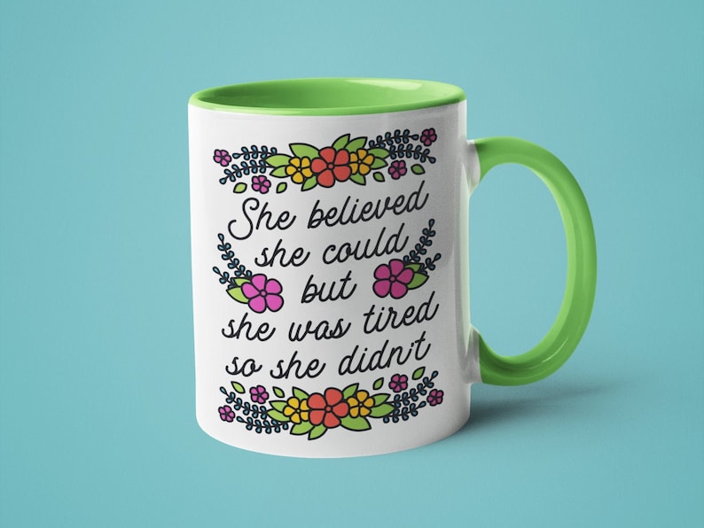 May include: White ceramic mug with a green rim and handle. The mug features a floral design and the text "She believed she could but she was tired so she didn't."