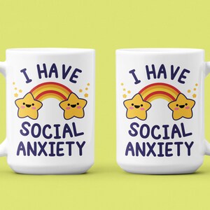 May include: Two white coffee mugs with a rainbow and two stars with faces. The text on the mugs reads "I have social anxiety".
