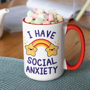 May include: White ceramic mug with a red handle. The mug has a rainbow and two yellow stars with faces on it. The text "I have social anxiety" is printed on the mug in purple.