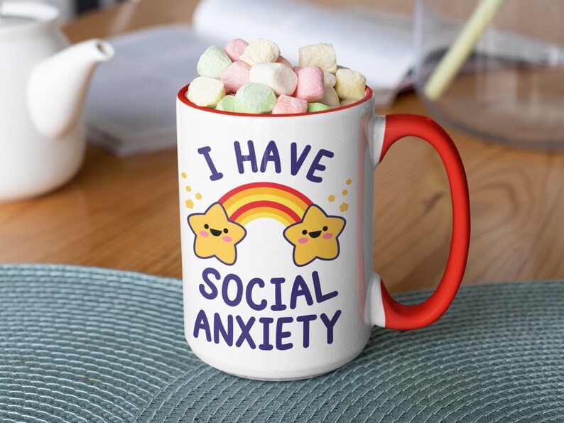 May include: White ceramic mug with a red handle. The mug has a rainbow and two yellow stars with faces on it. The text "I have social anxiety" is printed on the mug in purple.