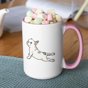 May include: White ceramic coffee mug with a pink handle. The mug features a cartoon illustration of a French bulldog doing yoga. The mug is filled with colorful marshmallows.
