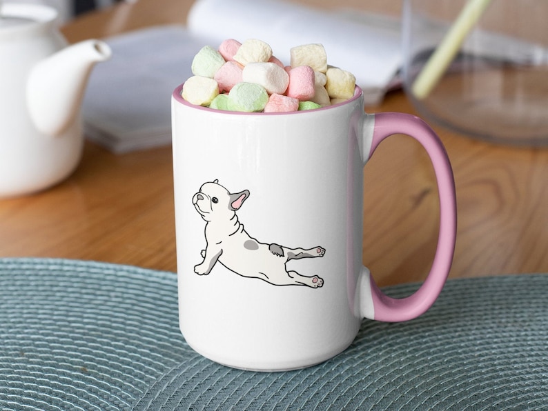 May include: White ceramic coffee mug with a pink handle. The mug features a cartoon illustration of a French bulldog doing yoga. The mug is filled with colorful marshmallows.