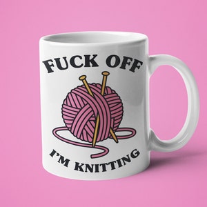 K&ouml;nnte beinhalten: Wei&szlig;e Keramiktasse mit einem schwarzen und rosa Motiv. Das Motiv sagt "F*ck Off I'm Knitting" und zeigt eine rosa Wollkn&auml;uel mit zwei Stricknadeln.