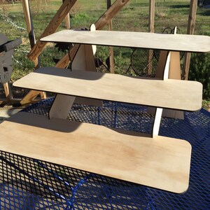Puede incluir: Un expositor de madera de tres niveles hecho de madera contrachapada. El expositor est&aacute; sin terminar y tiene un color de madera natural. El expositor est&aacute; sobre una mesa de metal azul.