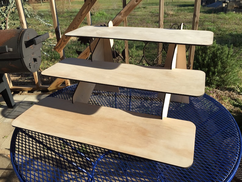 Puede incluir: Un expositor de madera de tres niveles hecho de madera contrachapada. El expositor est&aacute; sin terminar y tiene un color de madera natural. El expositor est&aacute; sobre una mesa de metal azul.