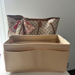 Op de afbeelding: Beige vilten handtas organizer met meerdere compartimenten en een ritssluiting aan de bovenkant. Een geruite tote bag met bruine leren randen en handvatten is zichtbaar op de achtergrond. Een kleurrijke sjaal is aan de handvat van de tote bag geknoopt.