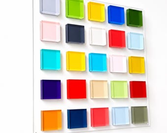 Puede incluir: Un panel de pared cuadrado que presenta una cuadrícula de veinticinco cuadrados acrílicos elevados, translúcidos y opacos en varios colores, incluidos rojo, azul, amarillo, verde, naranja, rosa, blanco y negro, dispuestos en cinco filas.