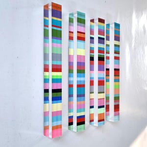 Peut inclure: Œuvre d'art abstraite murale composée de cinq panneaux verticaux en acrylique transparent. Chaque panneau est orné de rayures horizontales de différentes couleurs, dont le rose, le bleu, le vert, le jaune et le rouge. L'œuvre est fixée sur un mur blanc.