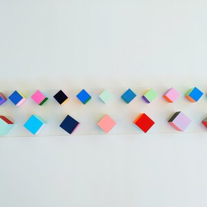 May include: A row of 18 colorful, geometric, wooden blocks arranged on a white surface. The blocks are painted in various shades of blue, green, pink, red, and black.