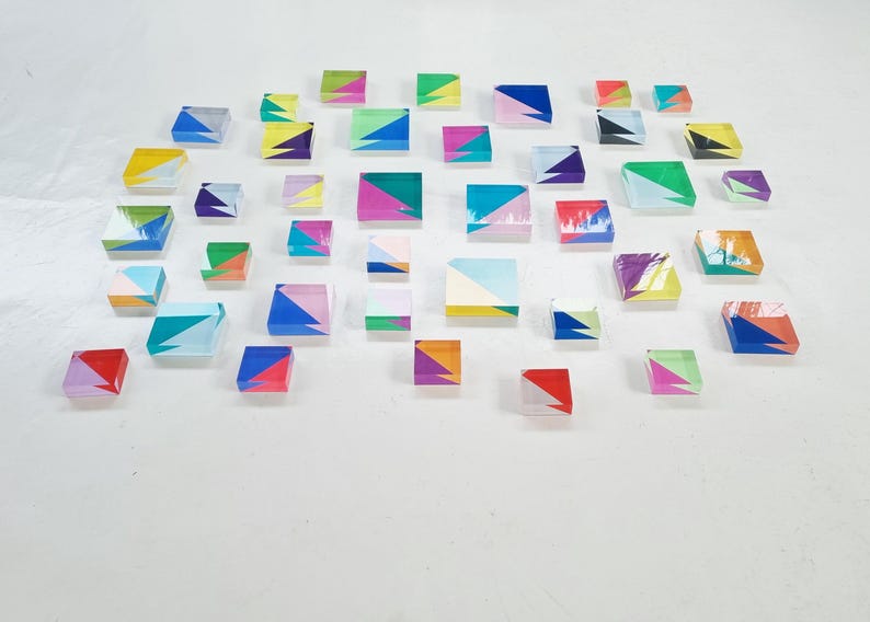 May include: A collection of colorful, geometrically patterned, square-shaped acrylic tiles. The tiles are arranged in a staggered pattern on a white surface.