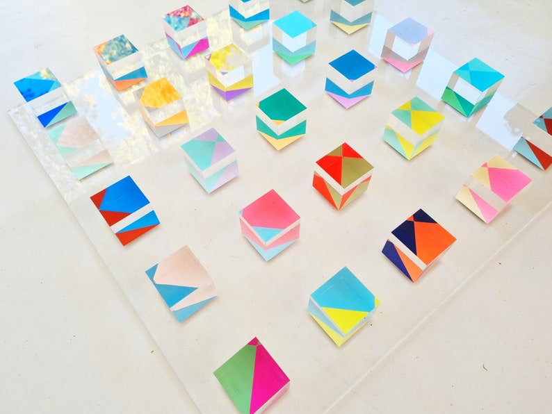 K&ouml;nnte beinhalten: Eine Sammlung von bunten, geometrisch gemusterten W&uuml;rfeln, die in einem Raster auf einer durchsichtigen Acryloberfl&auml;che angeordnet sind. Jeder W&uuml;rfel verf&uuml;gt &uuml;ber eine einzigartige Kombination aus Farben und Formen, die ein visuell anregendes und abstraktes Design schaffen.