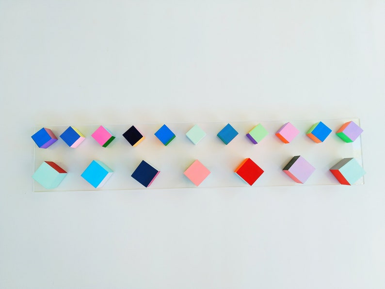 May include: A row of 18 colorful, geometric, wooden blocks arranged on a white surface. The blocks are painted in various shades of blue, green, pink, red, and black.