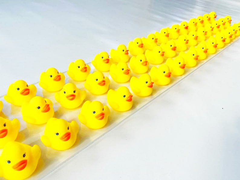 May include: A line of yellow rubber duck toys arranged in a row on a white surface. The ducks are all facing the same direction and have orange beaks and black eyes.