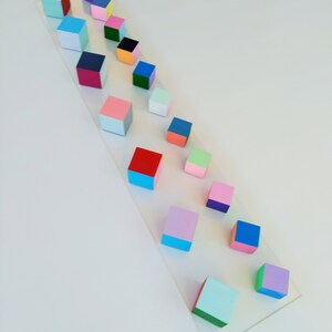 May include: A row of colorful wooden blocks arranged in a descending size order on a white surface. The blocks are painted in various colors, including red, blue, green, pink, and yellow.