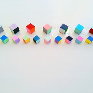 May include: A row of 18 colorful wooden cubes, each painted with two different colors, arranged on a clear acrylic surface. The cubes are in various shades of blue, green, pink, yellow, red, and gray.