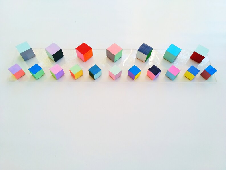 May include: A row of 18 colorful wooden cubes, each painted with two different colors, arranged on a clear acrylic surface. The cubes are in various shades of blue, green, pink, yellow, red, and gray.