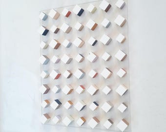 May include: Abstract wall art featuring a grid of white cubes with painted sides in various colors, including red, blue, and brown. The cubes are mounted on a clear acrylic sheet, creating a three-dimensional effect. The artwork is displayed against a white wall.