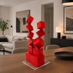 Op de afbeelding: Een moderne rode sculptuur bestaande uit gestapelde en schuine kubusvormen. De sculptuur staat op een heldere acrylbasis, geplaatst op een houten eettafel. De kamer heeft neutrale tinten, een bank en kunstwerken.
