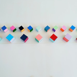 May include: A row of 16 small wooden cubes, each painted in two colors. The cubes are arranged in a line on a clear surface. The colors include pink, blue, green, red, purple, orange, black, and white.