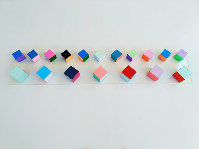 May include: A row of 16 small wooden cubes, each painted in two colors. The cubes are arranged in a line on a clear surface. The colors include pink, blue, green, red, purple, orange, black, and white.