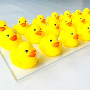 May include: A group of yellow rubber duckies lined up on a clear surface. The duckies have orange beaks and blue eyes.
