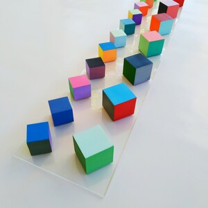 May include: A row of colorful wooden blocks arranged in a diagonal line on a white surface. The blocks are painted in various shades of blue, green, red, orange, yellow, pink, purple, and black.