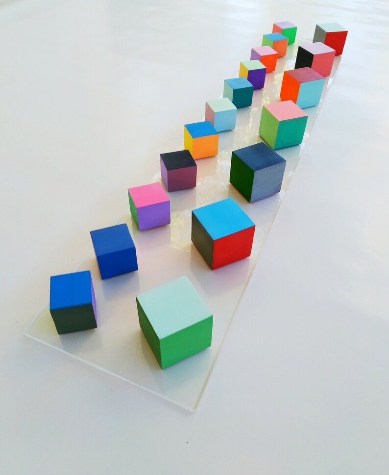 May include: A row of colorful wooden blocks arranged in a diagonal line on a white surface. The blocks are painted in various shades of blue, green, red, orange, yellow, pink, purple, and black.