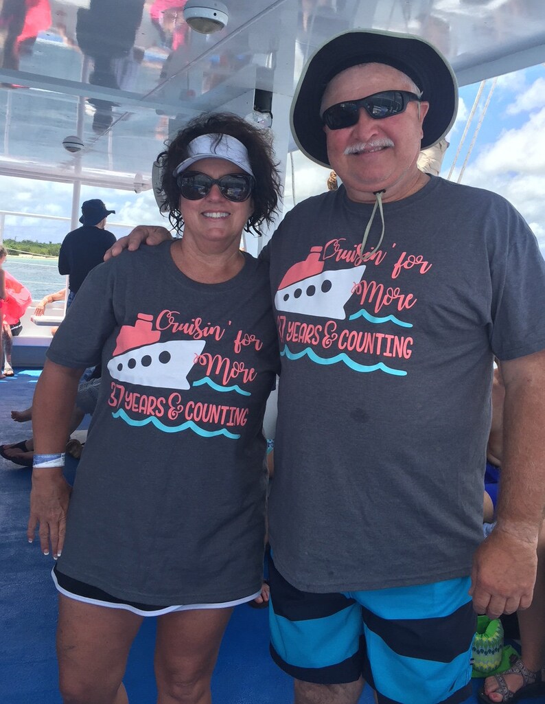 May include: A couple wearing matching gray t-shirts with a cruise ship graphic and the text "Cruisin' for More 37 Years & Counting". The man is wearing a black hat and sunglasses. The woman is wearing a white visor and sunglasses. They are standing on a boat.