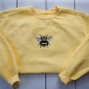 May include: A yellow sweatshirt with a crew neck and long sleeves. The sweatshirt features an embroidered bumblebee design on the front. The bee has black and yellow stripes and white wings. The cuffs and neckline are ribbed.