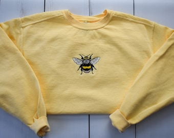 Könnte beinhalten: Ein gelbes Sweatshirt mit Rundhalsausschnitt und langen Ärmeln. Das Sweatshirt hat ein gesticktes Hummeldesign auf der Vorderseite. Die Biene hat schwarz-gelbe Streifen und weiße Flügel. Die Manschetten und der Halsausschnitt sind gerippt.