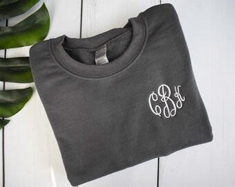 Può includere: Una felpa grigio antracite con un monogramma ricamato bianco. La felpa girocollo è piegata su una superficie di legno bianco, con una grande foglia verde nell'angolo in alto a sinistra. Il monogramma è in corsivo.