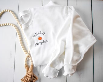 Könnte beinhalten: Ein weißes Sweatshirt mit dem gestickten Text "HELLO pumpkin" und einer orangefarbenen Kürbisgrafik. Das langärmlige Shirt ist gefaltet, die Manschetten sind sichtbar. Eine Holzperlen-Girlande mit braunen Quasten befindet sich im Hintergrund.