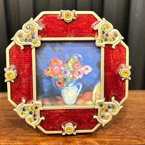 Op de afbeelding: Een rood achthoekig fotolijstje met gouden accenten en kleurrijke strass steentjes. Het lijstje heeft een kleine print van een stilleven met bloemen in een vaas.