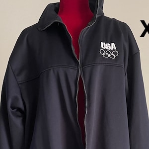 Peut inclure: Veste noire zippée avec un logo USA blanc et des anneaux olympiques sur le devant. La veste est de taille XXL.