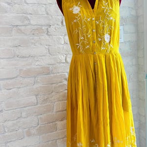 May include: A sleeveless, mustard yellow dress with white floral embroidery. The dress features a collared neckline, button-down front, and a pleated skirt. The dress is displayed on a mannequin.