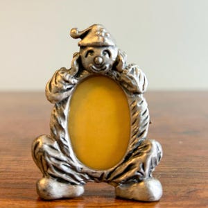 May include: A silver-colored, whimsical picture frame in the shape of a seated clown. The frame has an oval opening with a yellow background. The clown has a smiling face and a pointed hat. The frame is made of a metallic material.