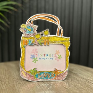 May include: A yellow and pink glitter picture frame shaped like a beach bag with a flower and sunglasses design. The frame says "Beach" and "SixTrees 3.5 inch x 5 inch".
