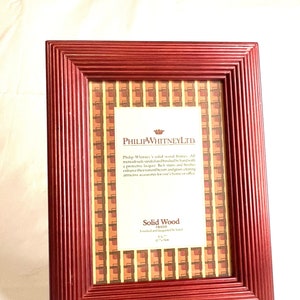 May include: A deep red wooden picture frame with a decorative patterned mount. The frame is labelled "Solid Wood Frame" and "Finished and lacquered by hand". The text "Philip Whitney Ltd." is also visible on the frame.