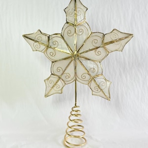 Peut inclure: Une étoile décorative pour sommet de sapin à cinq branches, composée de panneaux blancs translucides encadrés de métal doré. Chaque panneau présente des volutes en fil doré. L'étoile est fixée sur une base en spirale en métal doré.