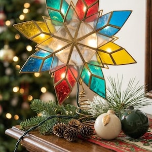 May include: A multi-pointed star tree topper featuring stained glass panels in red, blue, yellow, and teal, held together by a brass-toned metal frame. The star is illuminated from within and sits on a coiled metal base with a green electrical cord.