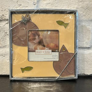 May include: A light yellow picture frame with a cat and fish design. The frame is 3x3 inches and 7x7 cm. The text "heart home" is printed on the frame. The frame is made by Connoisseur.