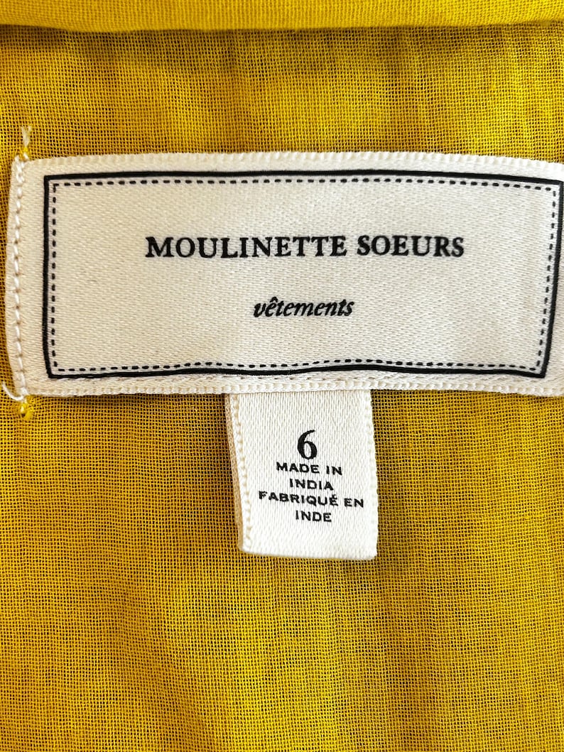 Puede incluir: Primer plano de una tela amarilla con una etiqueta blanca. La etiqueta dice "MOULINETTE SOEURS v&ecirc;tements" y "6 MADE IN INDIA FABRIQU&Eacute; EN INDE". La tela tiene un aspecto texturizado.