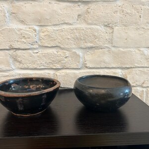 May include: Two small, dark ceramic bowls. The bowl on the left has a slightly irregular shape with a brown rim. The bowl on the right is rounder and has a smooth surface. Both bowls sit on a dark surface.
