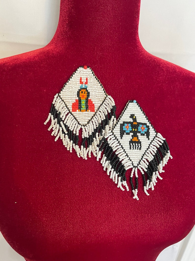 Puede incluir: Un par de pendientes de cuentas con un dise&ntilde;o nativo americano. Los pendientes son blancos con cuentas negras, rojas, azules y amarillas. Los pendientes tienen forma de diamante y tienen flecos.