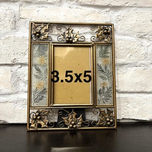 Op de afbeelding: Gouden metalen fotolijst met een opening van 3,5 x 5 inch. De lijst heeft een decoratief ontwerp van hulstbladeren en bessen. De lijst staat op een donker oppervlak.