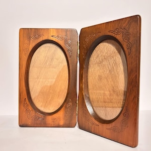 May include: A hinged, wooden picture frame with an oval opening. The frame is a warm brown color with decorative carvings in the corners. The frame is open, revealing the light-colored wood inside.