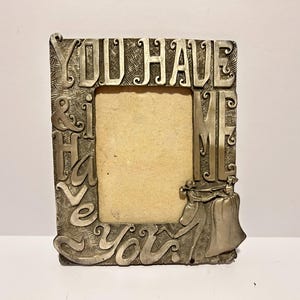 Puede incluir: Un marco de fotos plateado con un diseño en relieve de las palabras "You Have & I Have You" y una pequeña figura en la parte inferior derecha.